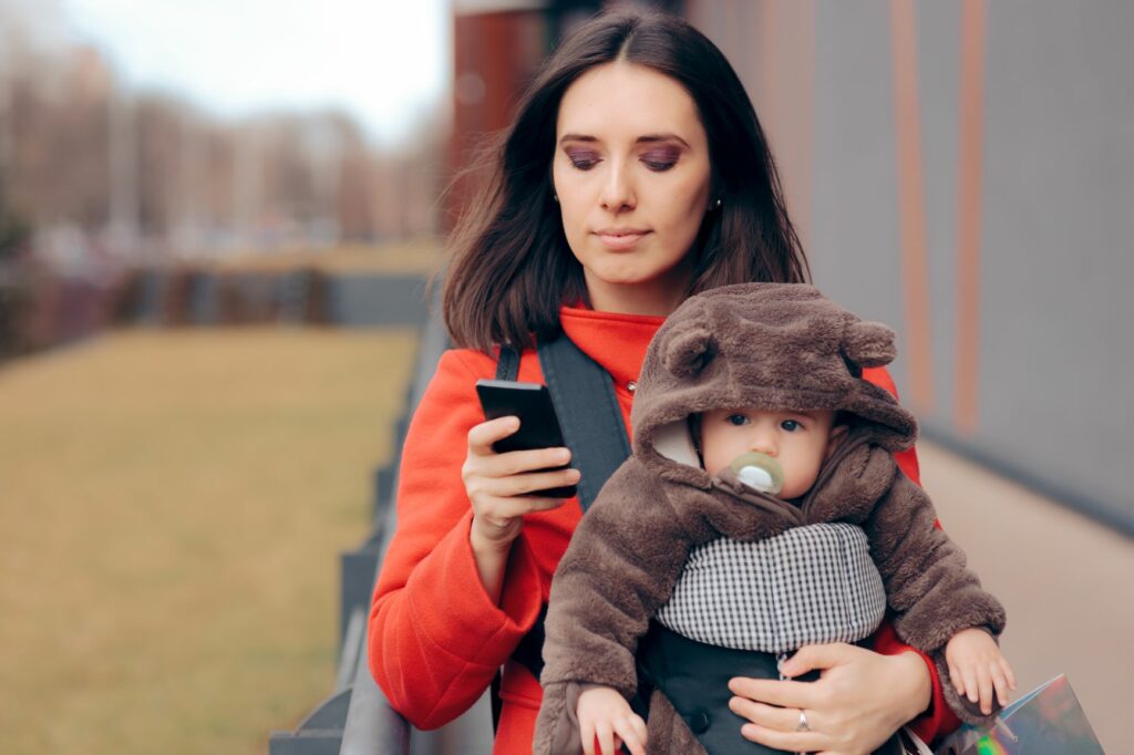 woman looking at her phone while walking with an infant in a bear hoodie rides along in a carrier