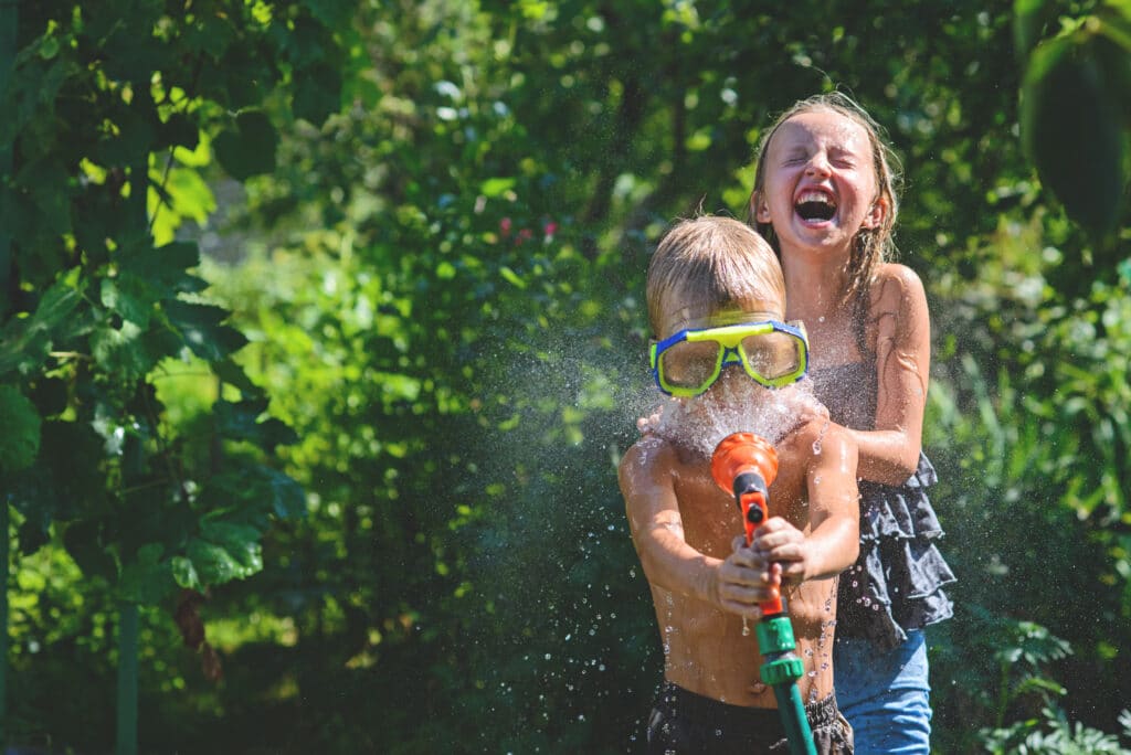 a little boy wearing goggles sprays himself and a girl behind him with water from a hose