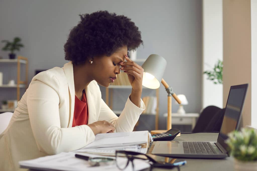 a woman looks stressed with with a pile of paperwork in front on her computer