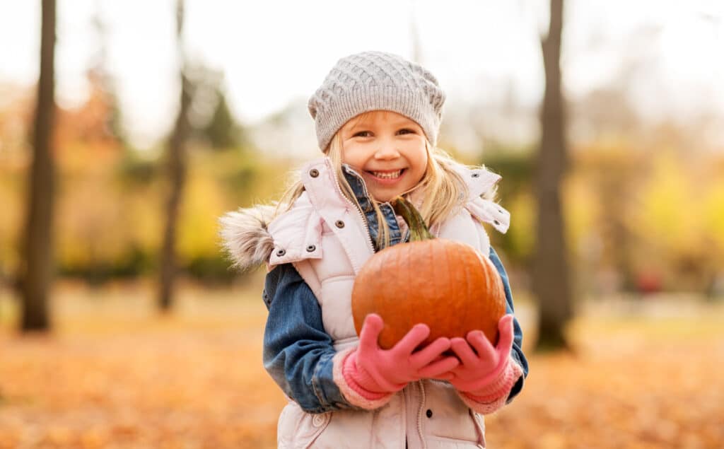 a young girl dressed for cold weather holds a pumpkin and grins at the camera