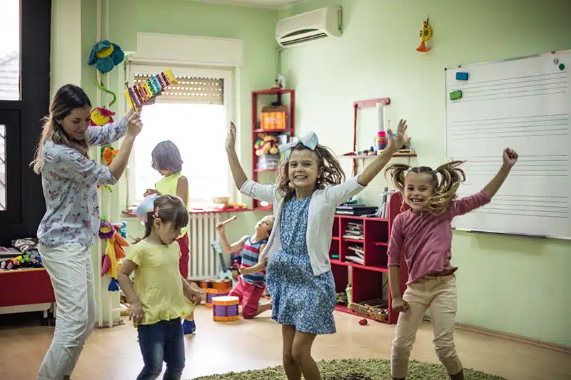 kids dance around their daycare classroom while a teacher plays a xylophone