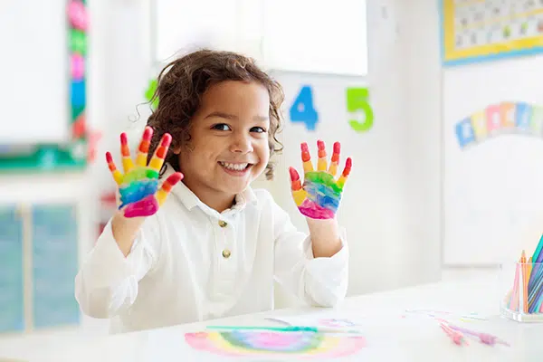 a preschooler smiles while showing rainbow-painted hands to the camera