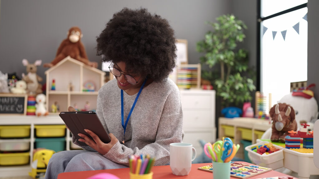 African american woman preschool teacher smiling confident writing on touchpad in a child care classroom.