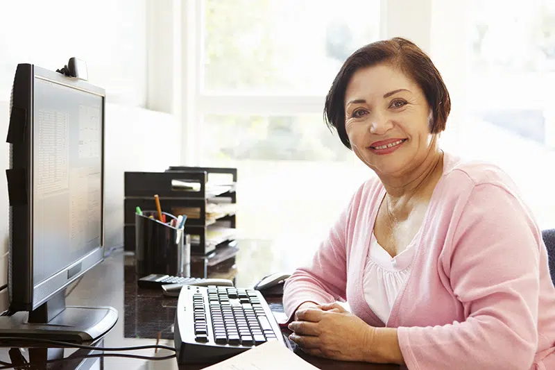 A middle aged woman sits in front of a computer and smiles sideways at the camera
