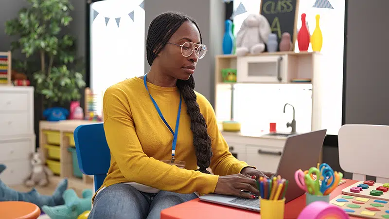 a woman types on a laptop computer in an empty child care classroom