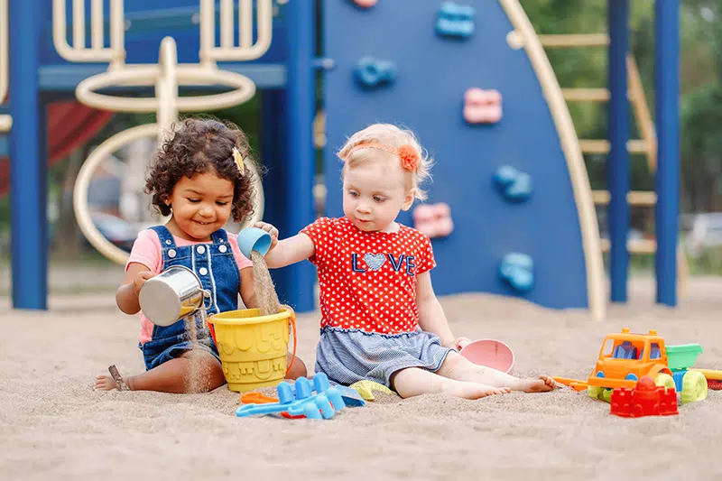 two toddlers at a playground pour sand into a yellow bucket