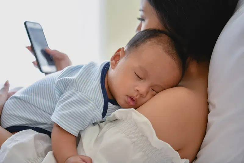 a woman does something on her phone while a baby is asleep in her chest
